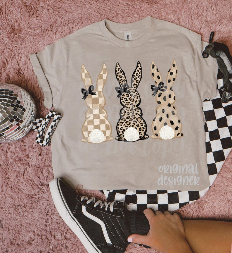 May include: A beige t-shirt featuring three bunny designs with checkered, leopard, and spotted patterns. Each bunny has a black bow and a white floral accent. The text "original designer" is printed on the shirt.