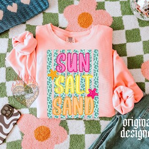 May include: A coral-colored sweatshirt with the words "SUN SALT SAND" in a colorful, playful font. The design includes a leopard print background and starfish accents. The words are in pink, yellow, and orange. The phrase "original designer" is printed in a script font.