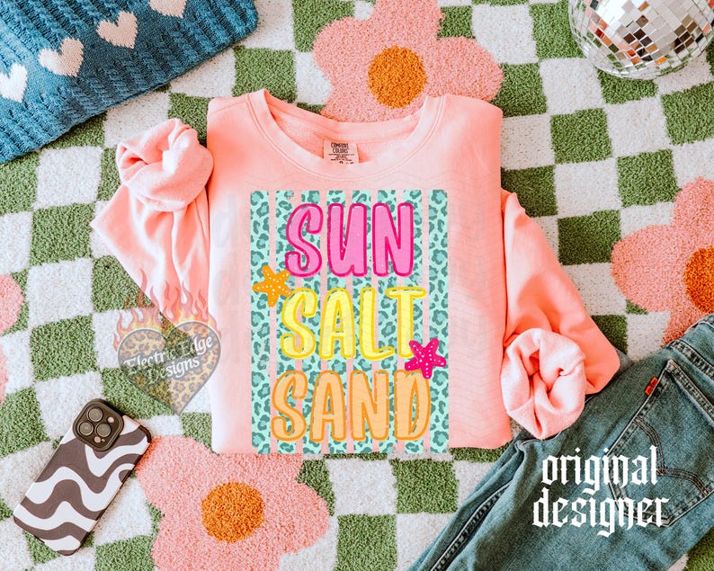May include: A coral-colored sweatshirt with the words "SUN SALT SAND" in a colorful, playful font. The design includes a leopard print background and starfish accents. The words are in pink, yellow, and orange. The phrase "original designer" is printed in a script font.