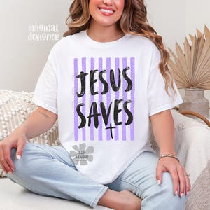 May include: White t-shirt with the text "JESUS SAVES" in black, set against a vertical striped background of purple and white. The shirt has a crew neck and short sleeves. The design includes a cross symbol.