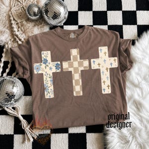 May include: A brown short-sleeve t-shirt featuring three graphic crosses across the chest. The left cross has a blue floral pattern, the center cross has a beige checkered pattern, and the right cross has a pattern of small blue and gold crosses.