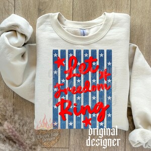May include: Cream-colored sweatshirt with the text "Let it Freedom Ring" in red script over a blue and white striped design. The design includes red stars. The sweatshirt has long sleeves and a crew neck.