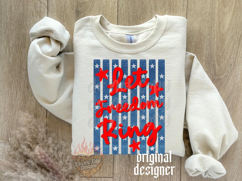 May include: Cream-colored sweatshirt with the text "Let it Freedom Ring" in red script over a blue and white striped design. The design includes red stars. The sweatshirt has long sleeves and a crew neck.