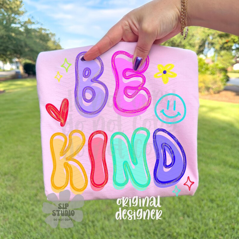 May include: A light pink sweatshirt with the words "BE KIND" in colorful bubble letters. The design includes a red heart, a yellow flower, a smiley face, and stars. The words "SIP STUDIO" and "original designer" are also printed on the sweatshirt.