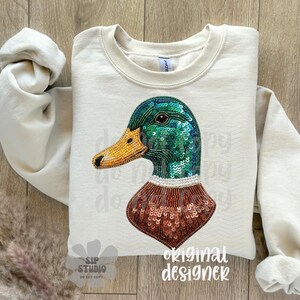 Pu&ograve; includere: Felpa color crema con un design dettagliato di un'anatra. La testa dell'anatra &egrave; adornata da paillettes verdi, un becco giallo perlato e un petto con paillettes marroni. Le maniche sono arrotolate e le parole "original designer" sono stampate in basso a destra.