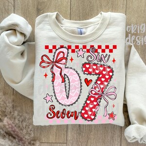 May include: Cream-colored sweatshirt with the numbers "6" and "7" in a heart-themed design. The number "6" is pink with white hearts, and the "7" is red with white hearts. The word "Six" and "Seven" are also printed on the shirt.