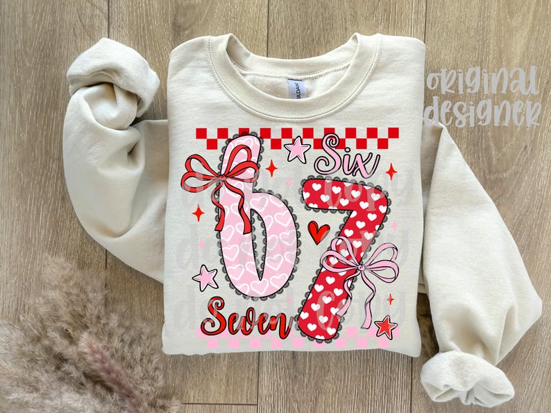 May include: Cream-colored sweatshirt with the numbers "6" and "7" in a heart-themed design. The number "6" is pink with white hearts, and the "7" is red with white hearts. The word "Six" and "Seven" are also printed on the shirt.