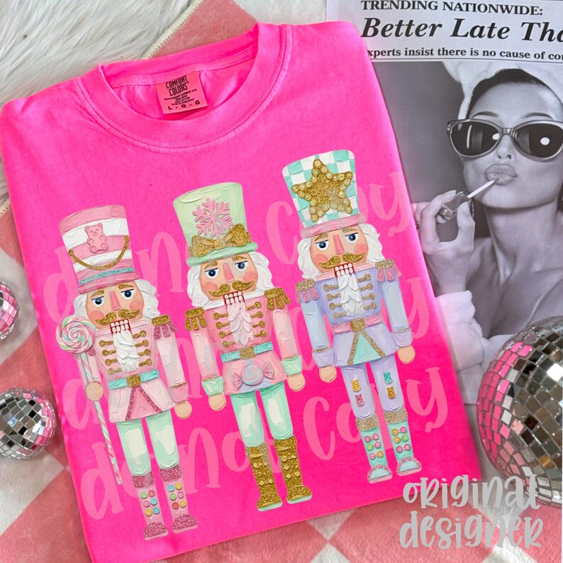 May include: A bright pink t-shirt featuring three colorful nutcracker illustrations. Each nutcracker has a unique design, with gold accents and pastel colors. The text "Candy Candy" is printed on the shirt. The brand label "Comfort Colors" is visible.