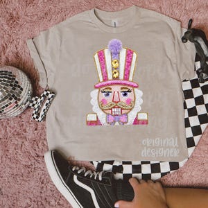 May include: A tan t-shirt featuring a colorful nutcracker design. The nutcracker has a pink and white striped hat, a pink bow tie, and a white beard. The text "original designer" is below the graphic.