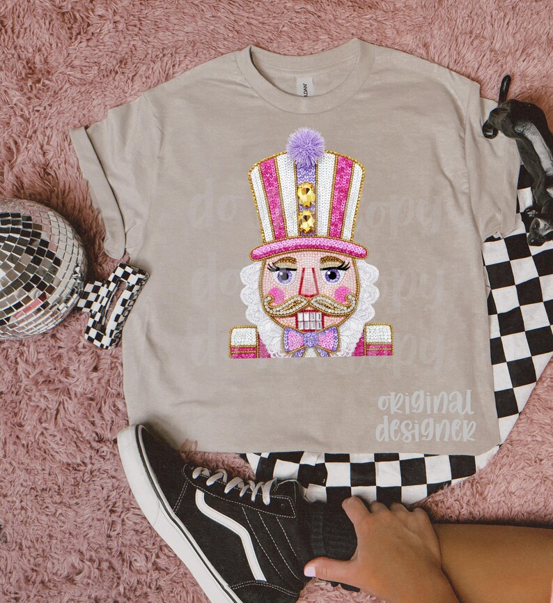 May include: A tan t-shirt featuring a colorful nutcracker design. The nutcracker has a pink and white striped hat, a pink bow tie, and a white beard. The text "original designer" is below the graphic.