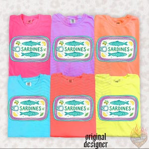 May include: Six colorful t-shirts, each with a unique hue, are displayed. Each shirt features a graphic of a sardine can with a fish illustration, the word "SARDINES", and decorative elements. The shirts are in shades of pink, purple, orange, blue, coral, and yellow.