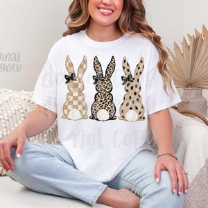May include: White t-shirt featuring three bunny designs with different patterns: checkered, leopard print, and spotted. Each bunny has a black bow. The words "original designer" are visible on the left side of the shirt.