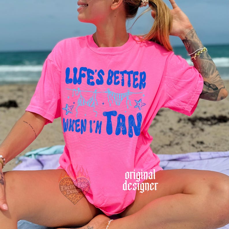 May include: A bright pink t-shirt with the text "LIFE'S BETTER WHEN I'M TAN" in blue. The design includes illustrations of swimwear and stars. The shirt is a casual, beach-themed apparel item.