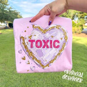 May include: A light pink sweatshirt featuring a large heart design with the word "TOXIC" in pink letters. The heart has a textured, painted appearance with gold glitter accents. Small heart details are scattered around the main design.