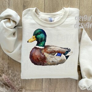 May include: Cream-colored sweatshirt featuring a detailed illustration of a mallard duck. The duck has a green head, yellow beak, and brown and blue body. The words "original designer" are visible on the top right.