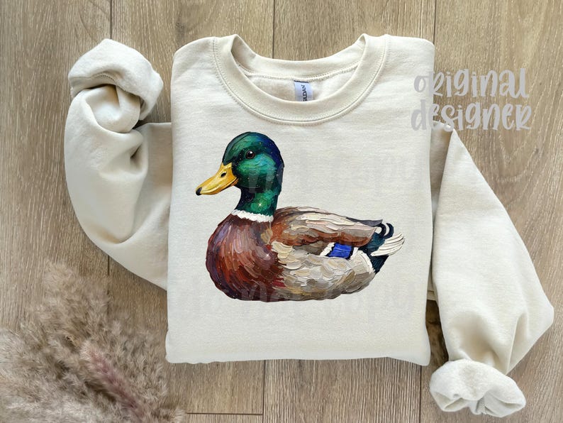 May include: Cream-colored sweatshirt featuring a detailed illustration of a mallard duck. The duck has a green head, yellow beak, and brown and blue body. The words "original designer" are visible on the top right.