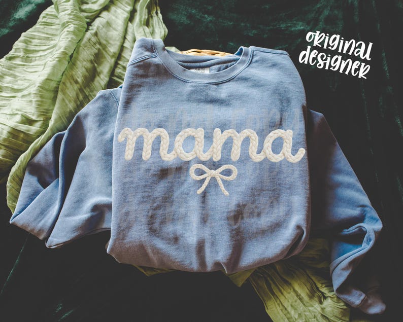 May include: A light blue sweatshirt with the word "mama" in white, rope-like lettering and a white bow. The words "original designer" are written in white script in the upper right corner. A green scarf is draped in the background.
