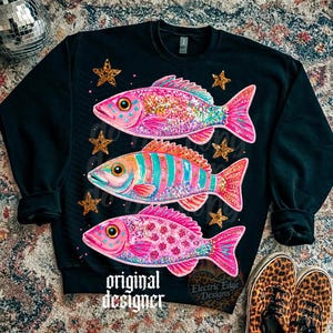 Puede incluir: Sudadera negra con tres dise&ntilde;os de peces coloridos con detalles brillantes y estrellas. Los peces son rosas, naranjas y azules. El texto "original designer" est&aacute; impreso en la parte inferior.