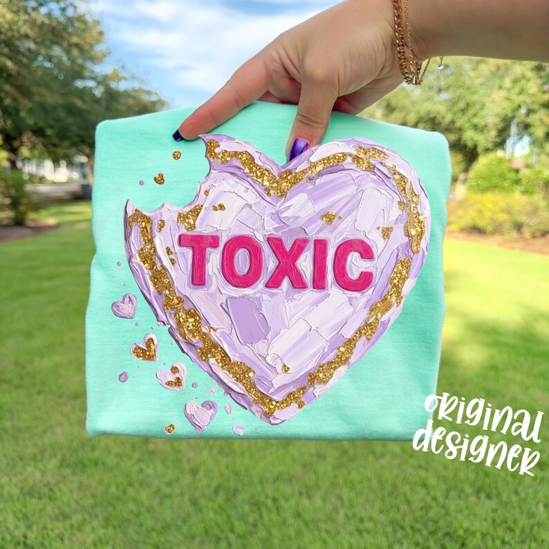 May include: A mint green t-shirt featuring a large, textured, purple heart with a bite taken out of it. The word "TOXIC" is in pink letters across the heart. Gold glitter accents the heart's edges and smaller hearts.