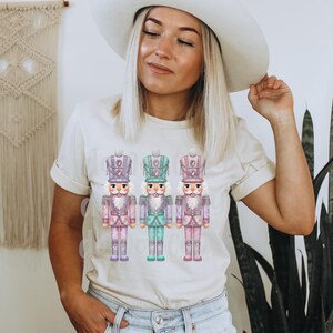 May include: A cream-coloured t-shirt featuring a watercolour illustration of three toy nutcracker soldiers. The nutcrackers are in pastel colours of pink, purple, and teal. The shirt is being worn by a person.