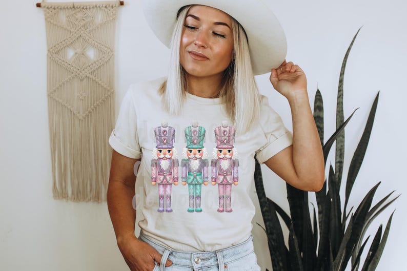 May include: A cream-coloured t-shirt featuring a watercolour illustration of three toy nutcracker soldiers. The nutcrackers are in pastel colours of pink, purple, and teal. The shirt is being worn by a person.