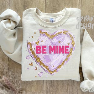 Op de afbeelding: Cr&egrave;mekleurige sweatshirt met een groot, getextureerd hartontwerp. Het hart is een mix van paars en goud, met de woorden "BE MINE" in roze. De mouwen zijn opgerold, op een houten achtergrond.