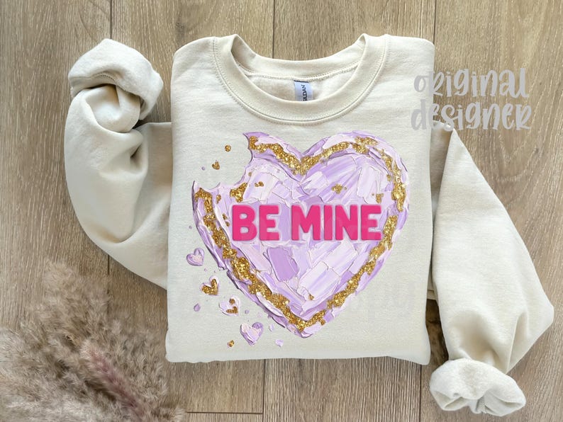 Op de afbeelding: Cr&egrave;mekleurige sweatshirt met een groot, getextureerd hartontwerp. Het hart is een mix van paars en goud, met de woorden "BE MINE" in roze. De mouwen zijn opgerold, op een houten achtergrond.