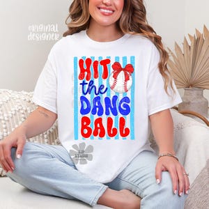 May include: White t-shirt with the text "Hit the Dang Ball" in red and blue, featuring a baseball with a red bow. The design is set against a backdrop of blue and white stripes. A casual, comfortable tee.