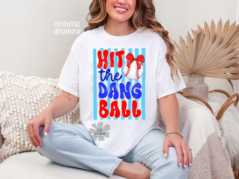 May include: White t-shirt with the text "Hit the Dang Ball" in red and blue, featuring a baseball with a red bow. The design is set against a backdrop of blue and white stripes. A casual, comfortable tee.