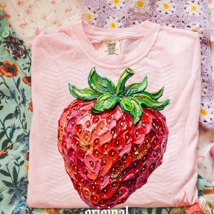 May include: A light pink t-shirt with a large, detailed strawberry graphic. The strawberry is red, pink, and gold, with green leaves. The text "original designer" is printed at the bottom. The shirt is from Comfort Colors.