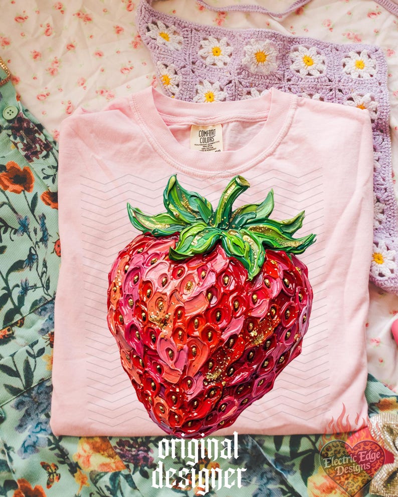 May include: A light pink t-shirt with a large, detailed strawberry graphic. The strawberry is red, pink, and gold, with green leaves. The text "original designer" is printed at the bottom. The shirt is from Comfort Colors.