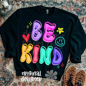 May include: Black sweatshirt with the words "BE KIND" in colorful bubble letters. The design includes a heart, a smiley face, stars, and a flower. The words "original designer" are printed at the bottom.