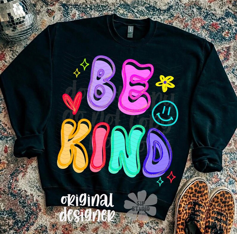 May include: Black sweatshirt with the words "BE KIND" in colorful bubble letters. The design includes a heart, a smiley face, stars, and a flower. The words "original designer" are printed at the bottom.
