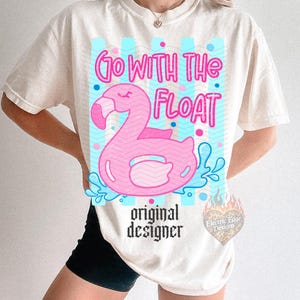 May include: Cream-colored t-shirt featuring a pink flamingo float with the text "Go With The Float" in pink. The design includes blue accents and the words "original designer" in black.