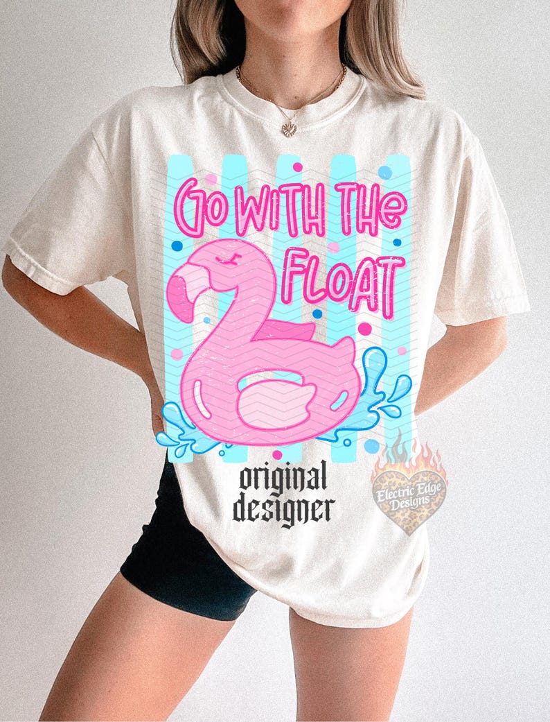 May include: Cream-colored t-shirt featuring a pink flamingo float with the text "Go With The Float" in pink. The design includes blue accents and the words "original designer" in black.