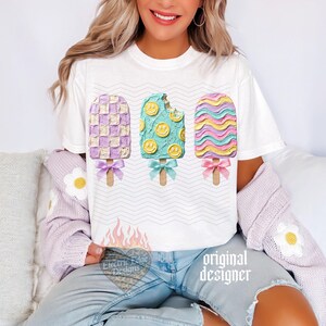 May include: A white short-sleeve t-shirt featuring a graphic of three popsicles with textured frosting. The left is purple and white checkered, the center is teal with yellow smiley faces, and the right has wavy pink, blue, and yellow stripes.