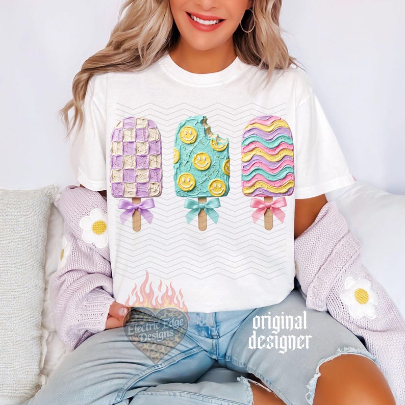 May include: A white short-sleeve t-shirt featuring a graphic of three popsicles with textured frosting. The left is purple and white checkered, the center is teal with yellow smiley faces, and the right has wavy pink, blue, and yellow stripes.