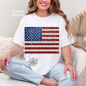May include: White t-shirt featuring a glittery American flag design. The flag has a blue square with white stars and red and white stripes. The shirt has short sleeves and a crew neck. The words "original designer" are printed on the top left.
