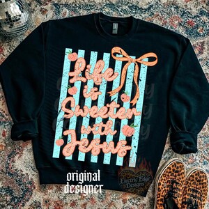 May include: Black sweatshirt with the text "Life is Sweeter with Jesus" in a cursive font, accented with peach-colored flowers and an orange bow. The design is set against a backdrop of light blue stripes. The words "original designer" are at the bottom.