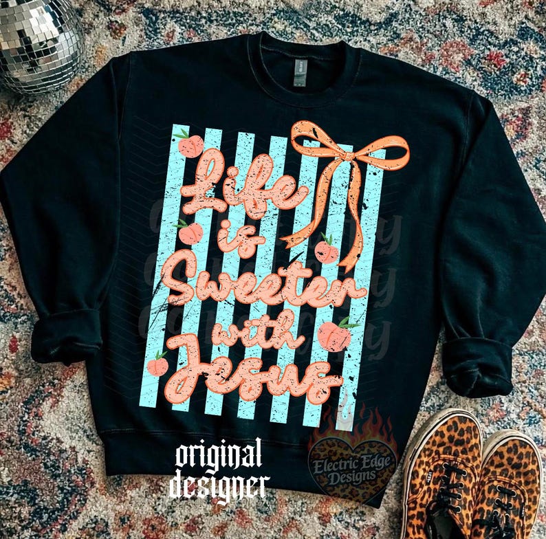 May include: Black sweatshirt with the text "Life is Sweeter with Jesus" in a cursive font, accented with peach-colored flowers and an orange bow. The design is set against a backdrop of light blue stripes. The words "original designer" are at the bottom.