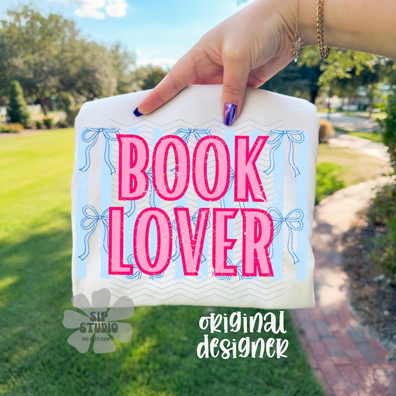 May include: White t-shirt with the words "BOOK LOVER" in pink, set against a light blue striped background with bow accents. The shirt also features the text "original designer".