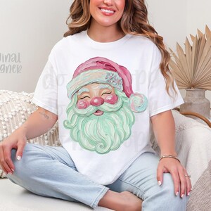 May include: White t-shirt featuring a watercolor illustration of Santa Claus. The Santa has a pink hat, rosy cheeks, and a mint green beard. The text "original designer" is visible on the left side of the shirt.