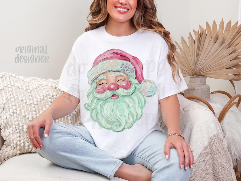 May include: White t-shirt featuring a watercolor illustration of Santa Claus. The Santa has a pink hat, rosy cheeks, and a mint green beard. The text "original designer" is visible on the left side of the shirt.