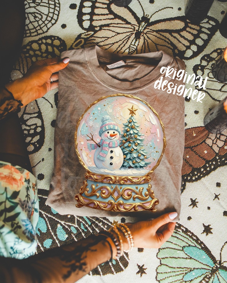 May include: A taupe t-shirt featuring a graphic of a snow globe containing a snowman wearing a blue hat and scarf next to a Christmas tree. The snow globe has an ornate gold base and the text original designer is printed in white script.