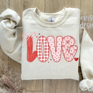 May include: A cream-colored sweatshirt with the word "LOVE" in a decorative design. The letters are red and pink, featuring gingham, hearts, and bows. A small red heart is next to the "E". The words "original designer" are in the upper right corner.