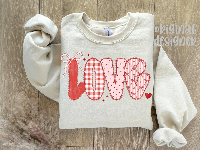 May include: A cream-colored sweatshirt with the word "LOVE" in a decorative design. The letters are red and pink, featuring gingham, hearts, and bows. A small red heart is next to the "E". The words "original designer" are in the upper right corner.