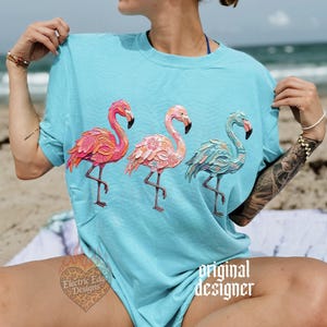 May include: A light blue t-shirt featuring three flamingo illustrations in shades of pink and teal. The flamingos are standing on one leg. The text "original designer" is printed on the shirt.