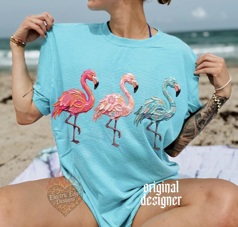 May include: A light blue t-shirt featuring three flamingo illustrations in shades of pink and teal. The flamingos are standing on one leg. The text "original designer" is printed on the shirt.
