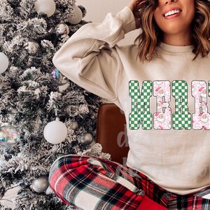 May include: A beige sweatshirt with the word "MERRY" in a green and white checkered pattern. The letters are outlined in black and filled with a Santa Claus and pink floral design. The person is wearing red and black plaid pants.