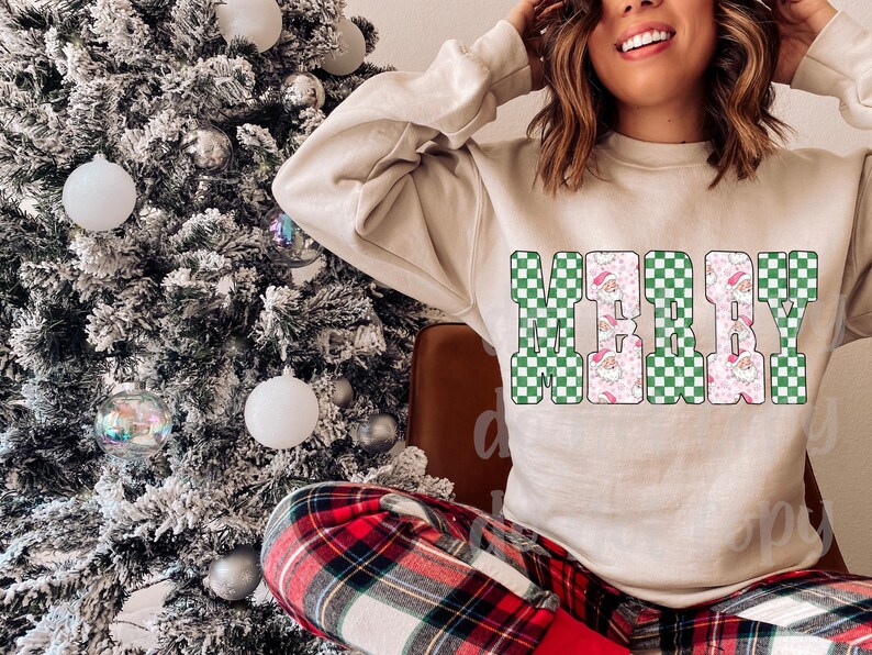 May include: A beige sweatshirt with the word "MERRY" in a green and white checkered pattern. The letters are outlined in black and filled with a Santa Claus and pink floral design. The person is wearing red and black plaid pants.